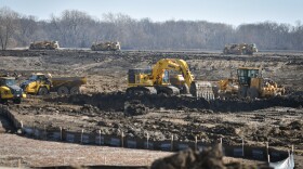 Exterior daytime image of an expansive dirt patch shows several yellow earth-moving vehicles at work.