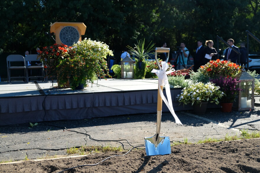 A silver and wood shovel with a white bow is stuck in the brown dirt in front of rises with color flowers around it. 
