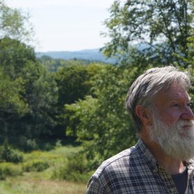 Man with gray hair and long gray beard, looking out into middle distance in front of verdant green background