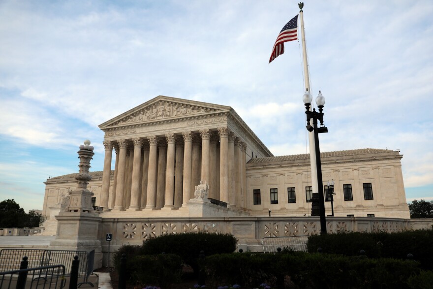 A large, faded white neoclassic style building looms in the center of the image. Several tall pillars hold up the center of the building. A red, white and blue American flag can also be seen flying in the foreground, to the right of the image. 