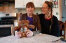 Two women sit at a kitchen table with a teddy bear, smiling