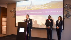 Governor Gretchen Whitmer and Lieutenant Governor Garlin Gilchrist stand at a stage in front of a large projector screen. The screen says: "Michigan Taiwan Office Opening Reception, Monday March 4, 2024." On the stage there is also a wooden podium. K.C Kong is standing by the podium speaking to the attendees. 