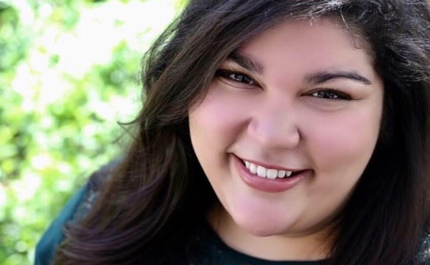 A smiling young woman with long, dark hair  is standing outside wearing a green shirt.