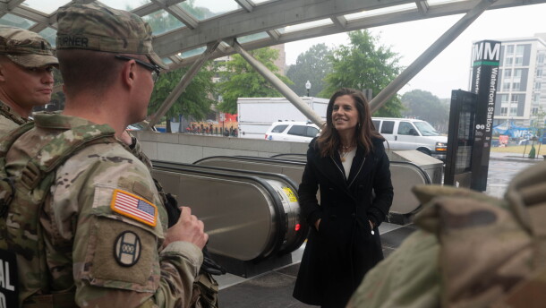 A woman in a black overcoat talks with soldiers in camouflage uniforms in a subway station. 