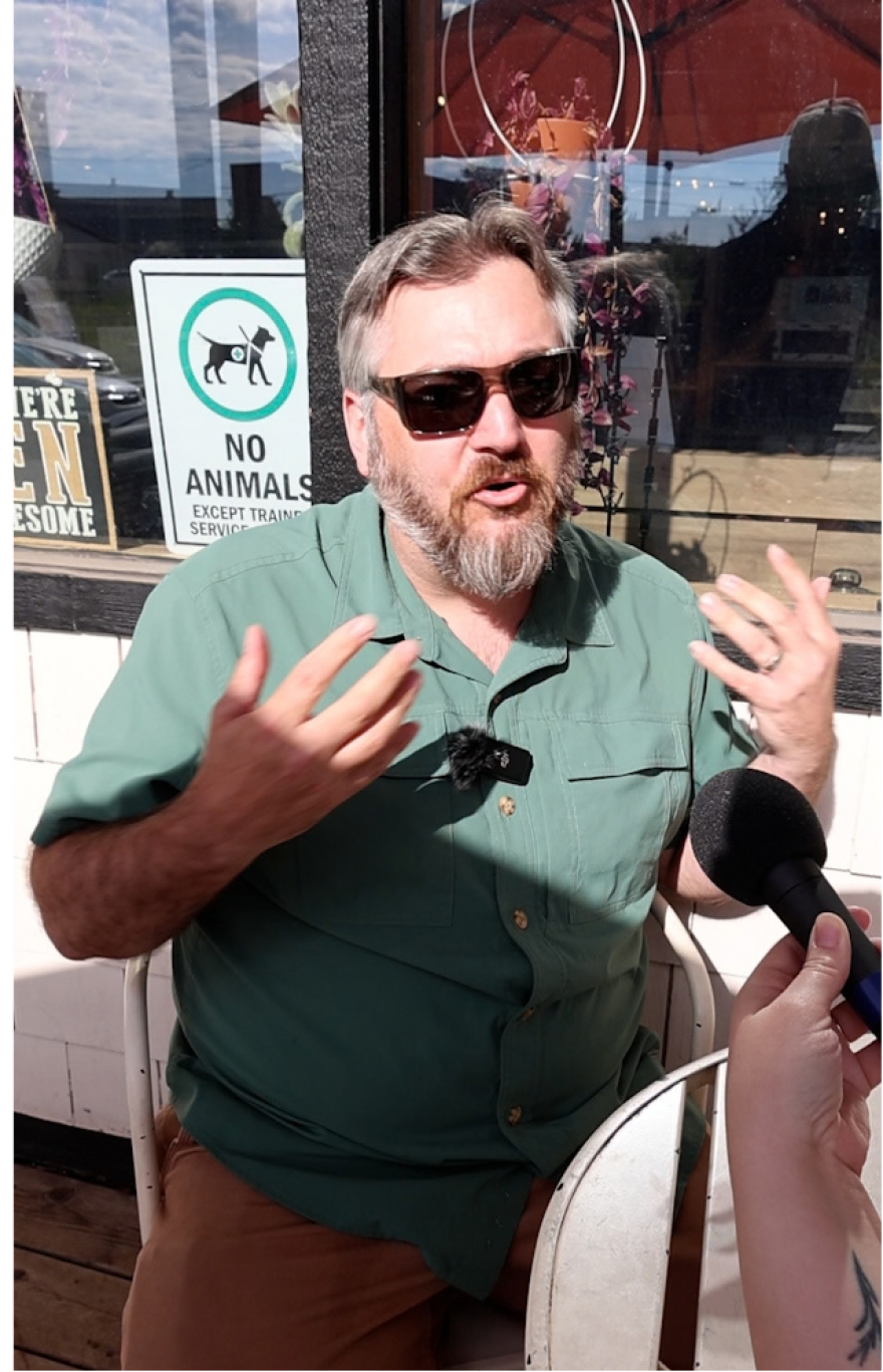 A white man in a green shirt and brown pants sits at a white cafe table. He has a salt and pepper beard and hair and is wearing sunglasses. He is talking into a microphone and gesticulating with his hands.