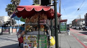 A woman stands next to her fruit cart on the corner of 16th and Mission. She has a big umbrella covering the cart to provide shade, and it is bursting with melons, oranges, papayas, guavas, and other fruits. 