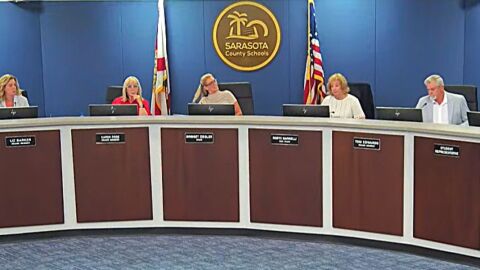 government dais with 4 women and 1 man sitting. behind them is a largo circle logo that says Sarasota County Schools, and a Florida and US Flag on poles 