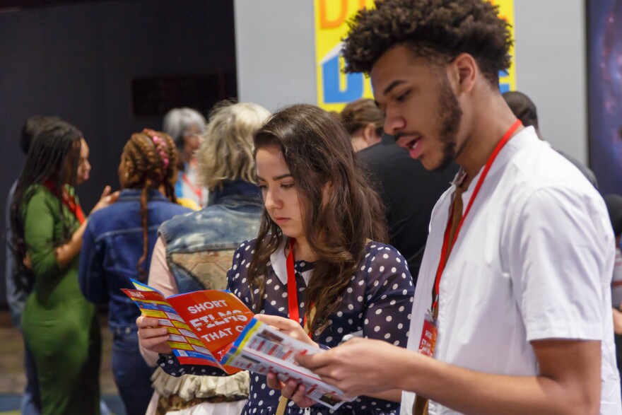 Two young people open brochures and look inside to see a list of films. In the background, a crowd lines up outside an auditorium.