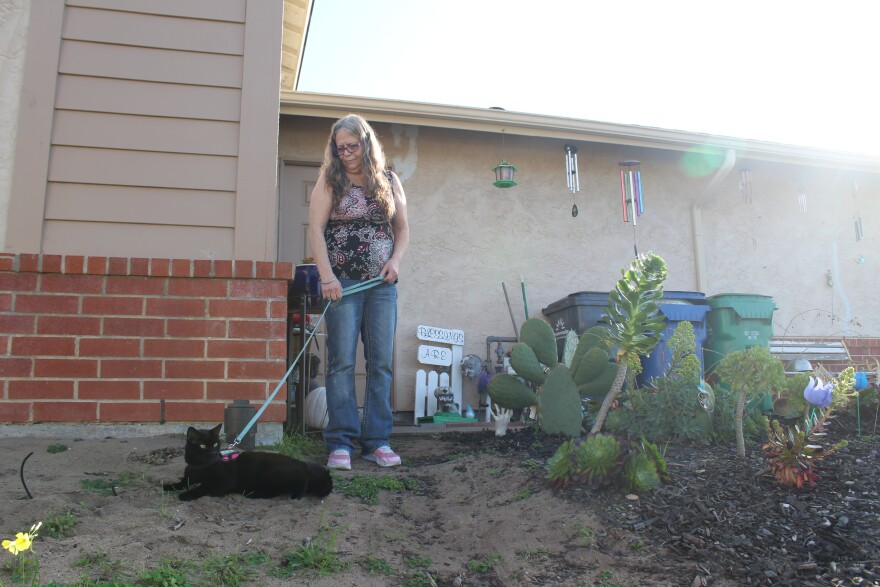 A woman wearing jeans and a patterned tank top holds a black cat on a leash in a patch of sandy dirt in front of a beige house with brick trim.