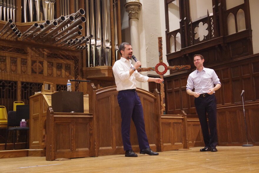 Pete Buttigieg speaks into a microphone and points at Sean McCann, who is standing next to him, on a wooden church stage in front of a large pipe organ. Both are wearing light button-up shirts and dark pants. McCann is looking at Buttigieg and smiling. 