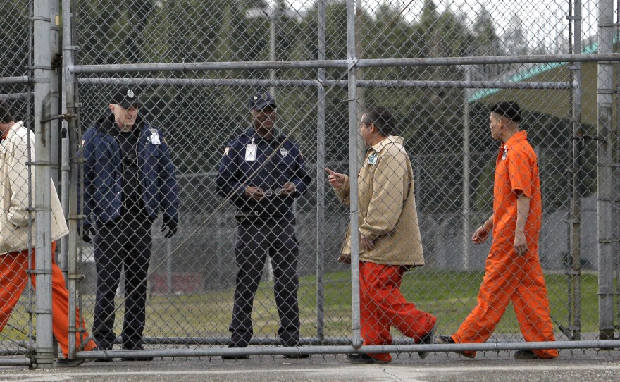 Men in orange jumpsuits and brown jackets walk past two men dressed in dark blue uniforms behind a chain-linked fence.