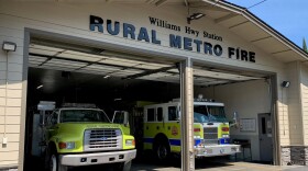 A fire station with two yellow/green fire trucks parked in a garage. The sign above says "Williams Hwy Station, Rural Metro Fire."