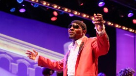 Black man wearing sunglasses and peach suit jacket singing on stage with his arms out