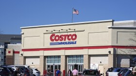 A tan building with American flag on the roof of it with a sign above the store that says, "Costco." People walking with baskets and driving in the parking lot. 