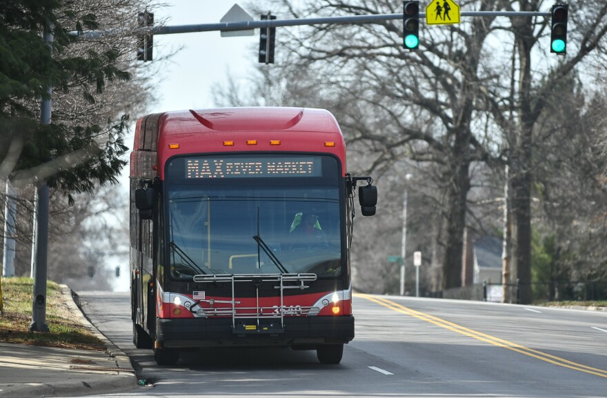 A red commuter bus moves on a quiet city street. It has a lighted sign on it that reads "MAX River Market."