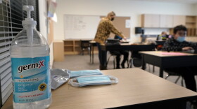 Hand sanitizer, wipes, and surgical masks rest on a desk in a classroom.