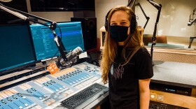 KUOW producer Kelsey Kupferer stands in a studio in front of a mic and two monitors. She has long blond hair and is wearing a black COVID safety mask. She's smiling to the camera.