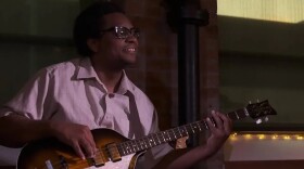A man playing a bass guitar, wearing glasses and smiling in a dimly lit interior space