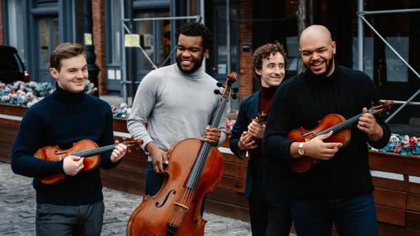 All four men wear dark jeans and turtlenecks. They smile and laugh together on a street in New York City, all while carrying their instruments.