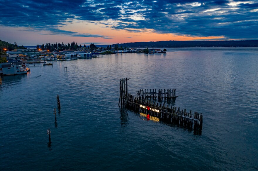 The "You'll Love Tacoma" sign — a temporary, guerrilla art installation at Titlow Beach in Tacoma — reflects on the water below during a summer sunset.