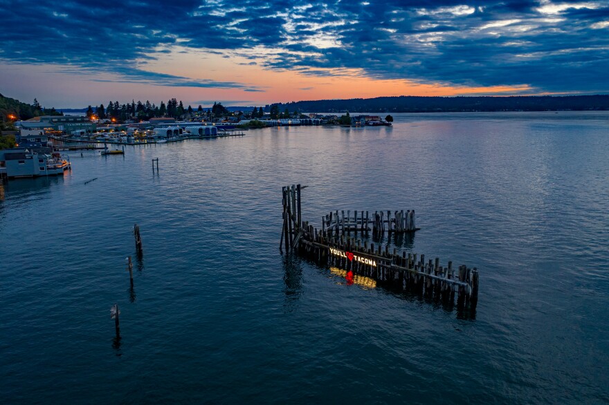 The "You'll Love Tacoma" sign — a temporary, guerrilla art installation at Titlow Beach in Tacoma — reflects on the water below during a summer sunset. 