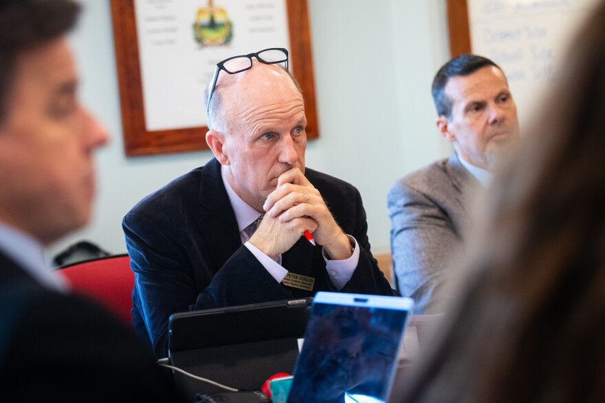 A white man in a suit with glasses pushed onto the top of his head sits in a room of other people that can't be clearly seen.