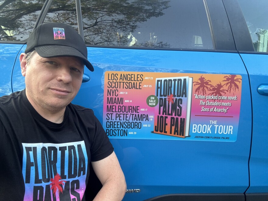 A man wearing a baseball cap sits in front of car with a sign about his book called 'Florida Palms.'