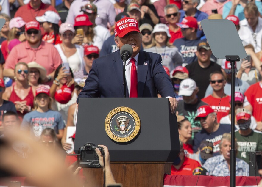 President Trump, wearing a blue suit, red tie and red ball cap talks at a podium bearing the presidential seal against a backdrop of people on a sunny day