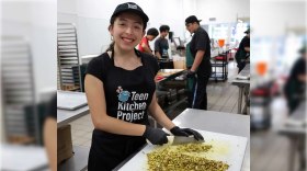 A smiling young woman with a baseball cap and apron wears black food-service gloves as she chops a pile of something on a white cutting board. Behind her more young adults work in a commercial kitchen filled with stainless steel counters and pans. 