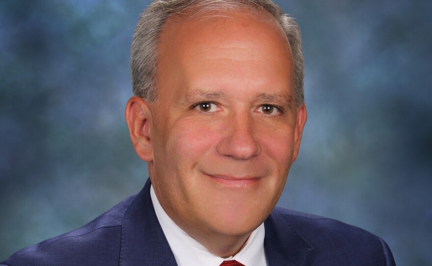 A man with short grey hair is wearing a navy blazer, red tie and white button-down shirt.