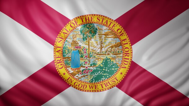 3d rendering of a detailed Florida flag, whihc is white, with red stripes coming down from the corners into an x pattern. The seal in the middle is yellow on the outside rim and says great seal of this state of florida, in god we trust. The illustration inside shows a woman in a long blue skirt depositing red and white followers into the water. There's a ship and palm tree in the background. 