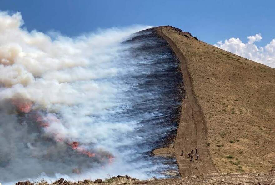 one half of a hillside is on fire, with winds pushing the fire and smoke down the hill