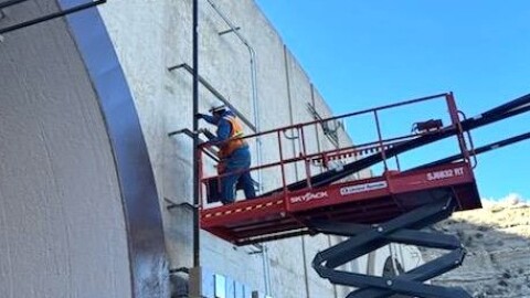 A person stands on a massive hydraulic lift as they work on some electrical wires on the side of a very tall tunnel.