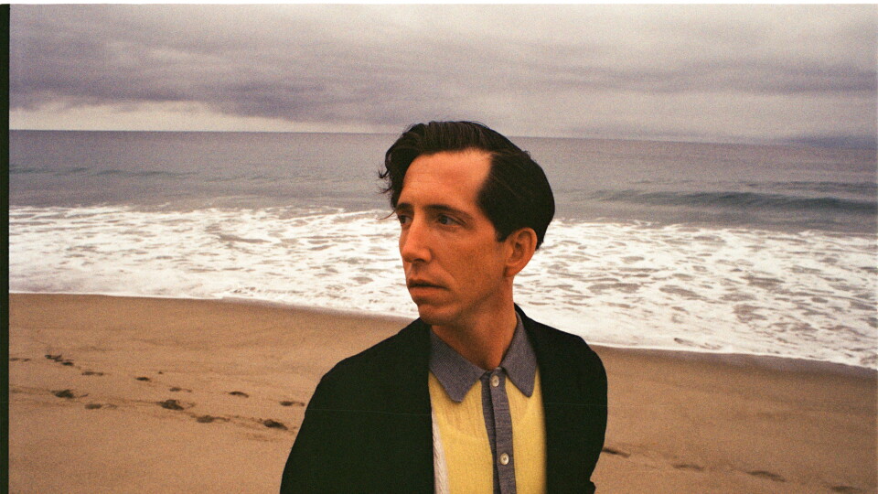 Pokey LaFarge looks off camera into the distance on a beach with the ocean behind him.