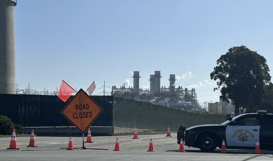 A road closed sign, police car, and traffic cones are shown in front of industrial buildings with smoke rising from them.
