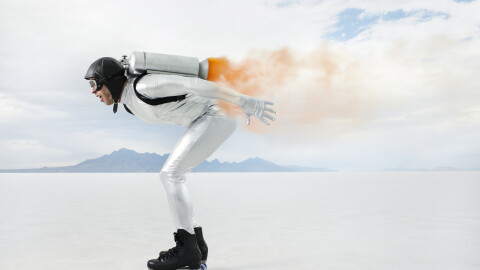 FILE: A wide, horizontal shot of a person wearing a white jumpsuit, black roller skates, and a jetpack, gliding across a vast, flat salt plain with mountains in the background under a cloudy sky. The person is in a crouched position with arms outstretched, surrounded by a cloud of orange smoke emitted from the jetpack. The scene is bathed in soft, diffused daylight, capturing a sense of adventure and innovation.