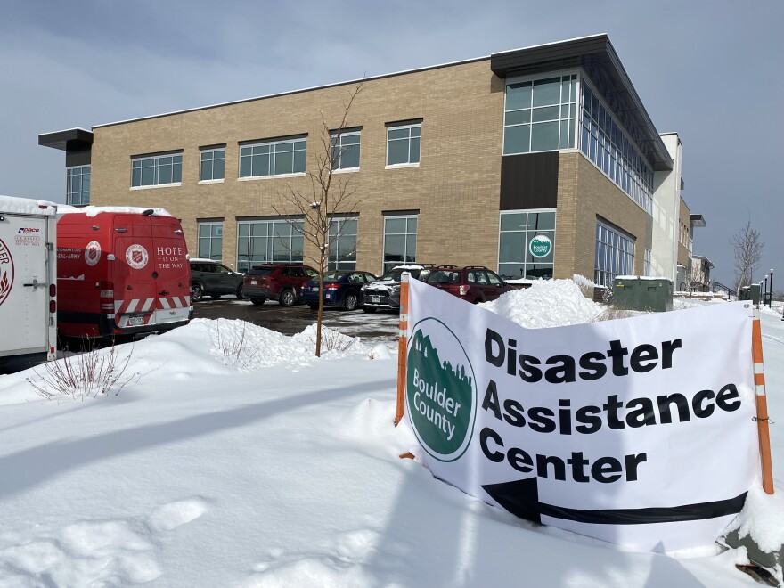 A sign that reads "Boulder County Disaster Assistance Center" stands in the snow in front of a parking lot and a two-story building.