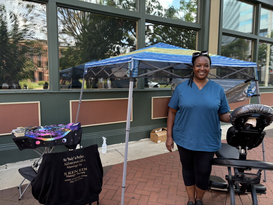 Woman in blue top and dark pants smiles for a portrait while standing under a pop-up awning, next to a massage chair