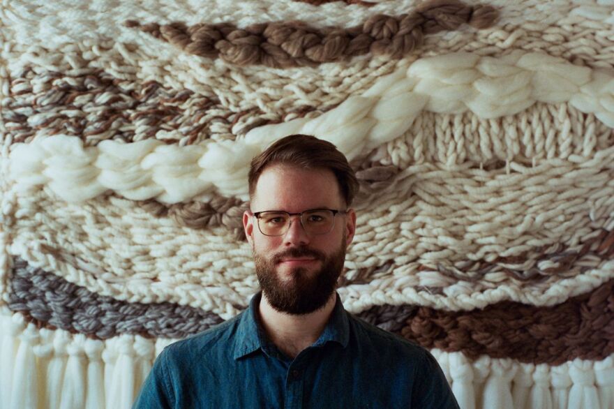 A man with glasses, a beard and blue shirt. In the background a wall hanging made of wool-like fabrics