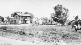 A black and white photo of a white farmhouse and a man on a horse. 