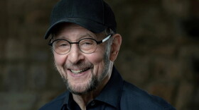 An older man in a ball cap, glasses and a dark blue button-down shirt smiles at the camera. 