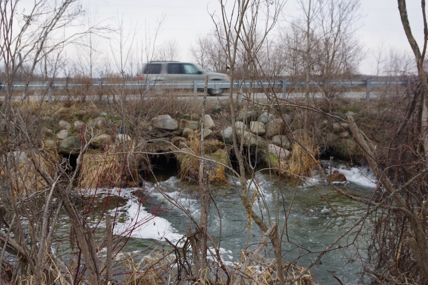 A light colored mini-van passes over a failing culvert on S. Patterson Road near Gun Lake.  A metal four-pipe culvert isn't large enough to handle the currents in the Gun River at low water levels, eroding the banks and making a fish passage barrier for the trout downstream.  