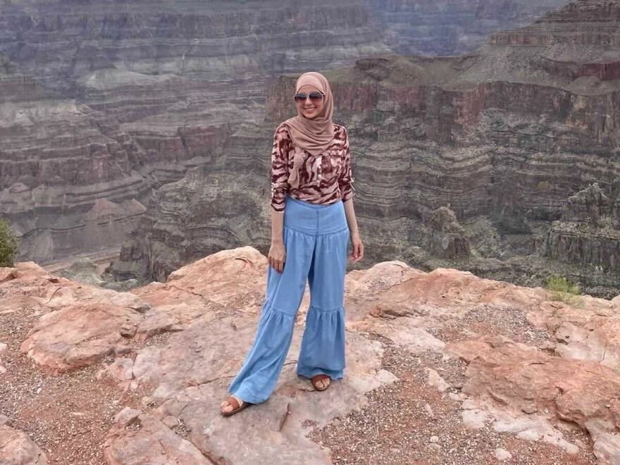 A woman wearing a  light rose colored headscarf, pink and brown blouse, blue pants and sandals poses for a photo at the Grand Canyon. 