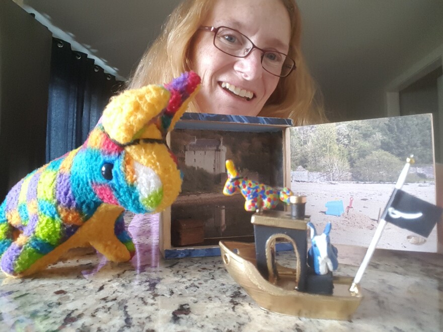 Photograph of artist with her diorama of handmaid ships and stuffed animals in front of pictures of Scottish castles. 