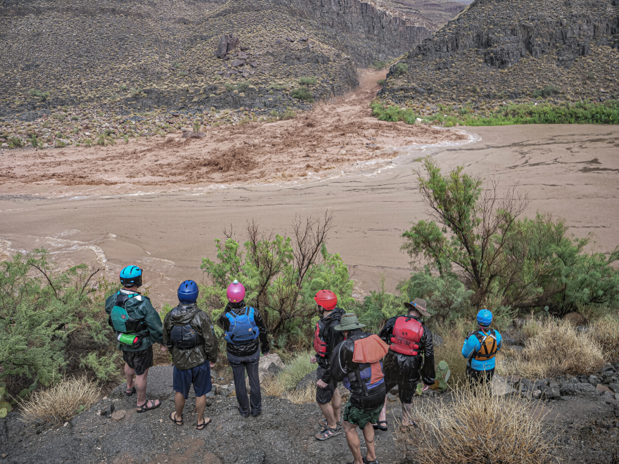 Rafters watch safely on the shore as a flash flood moves into the river from a canyon. The water is brown and rushing.