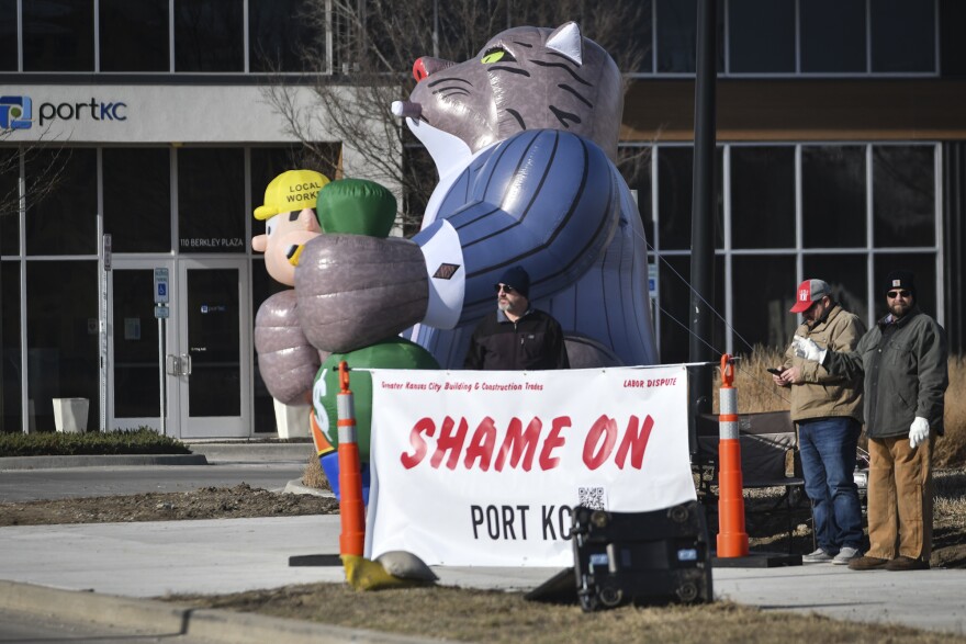 Three men can be seen standing outdoors on a corner of an intersection. They are standing behind a banner that reads "Shame on Port KC." Behind them is a large inflatable cat that is is smoking a cigar wearing a suite. It  has its paws wrapped around an inflatable person who looks like a construction worker in its right paw and a bag of money in its left paw.