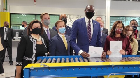 Garlin Gilchrist stands at the center of a group wearing suits and face masks behind blue-and-yellow industrial equipment. Fay Beydoun stands at front left. Gretchen Whitmer stands at front right holding a document. Several other people in suits stand behind them indoors.