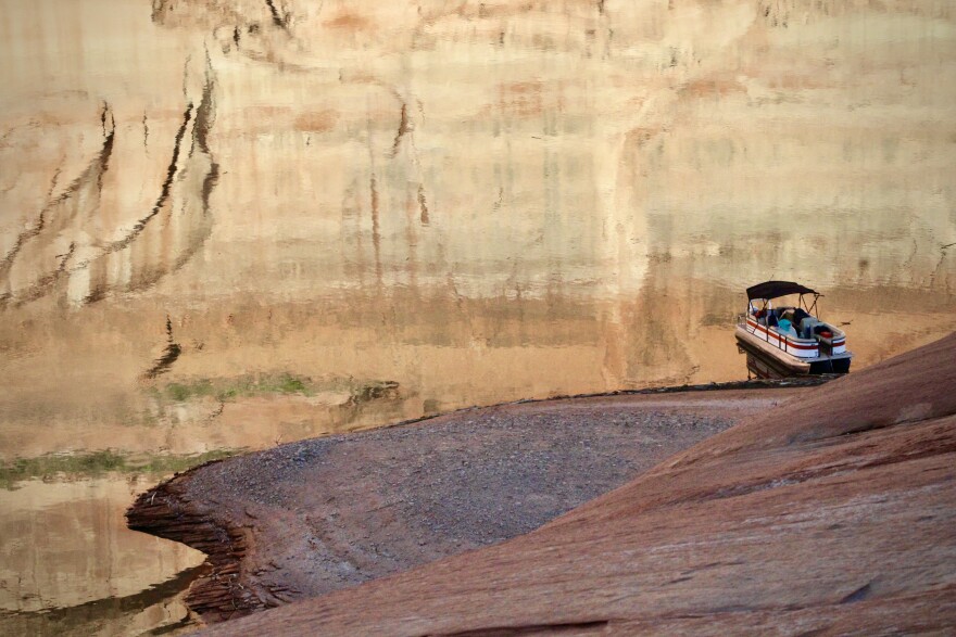 A pontoon boat is tied up at the shore of a recently-revealed beach in one of Lake Powell's side canyons on April 10, 2023. The evening sunlight casts a reflection of the canyons "bathtub rings" on the still water.