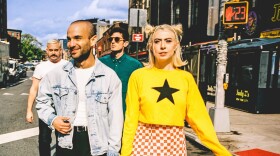  A woman and three men walk across a street in a city with shops on either side of the street.