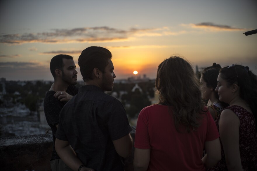 Group of men and women talk in a semi-circle. The sun is setting over a city behind them.